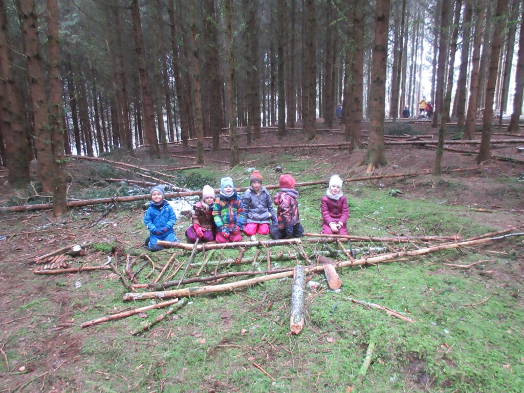 Gruppe von Kindern in einem Wald mit Holzstämmen und Stroh auf dem Boden.