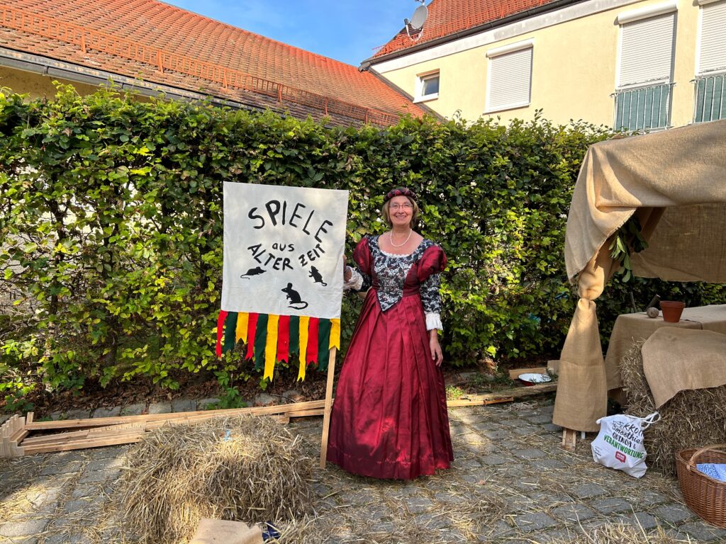 Mädchen in Altertum-Kleidung steht vor einem Banner mit der Aufschrift „SPIELE AUS ALTERZEIT“.