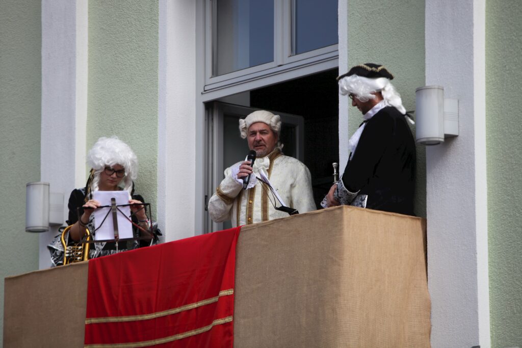 Männer und Frauen im historischen Kostüm auf der Veranda, vor einem roten Vorhang.