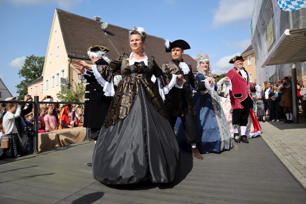 Eine Frau in historischer Kostüme führt eine Tanzveranstaltung auf einer erhöhten Plattform vor einer Menschenmenge durch.