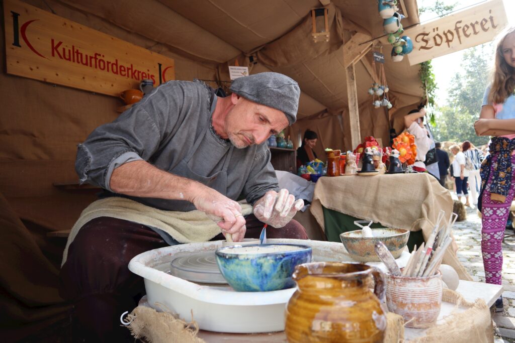 Maler am Keramikofen, im Hintergrund eine Person und ein Logo, Text „Kulturförderkreis“.