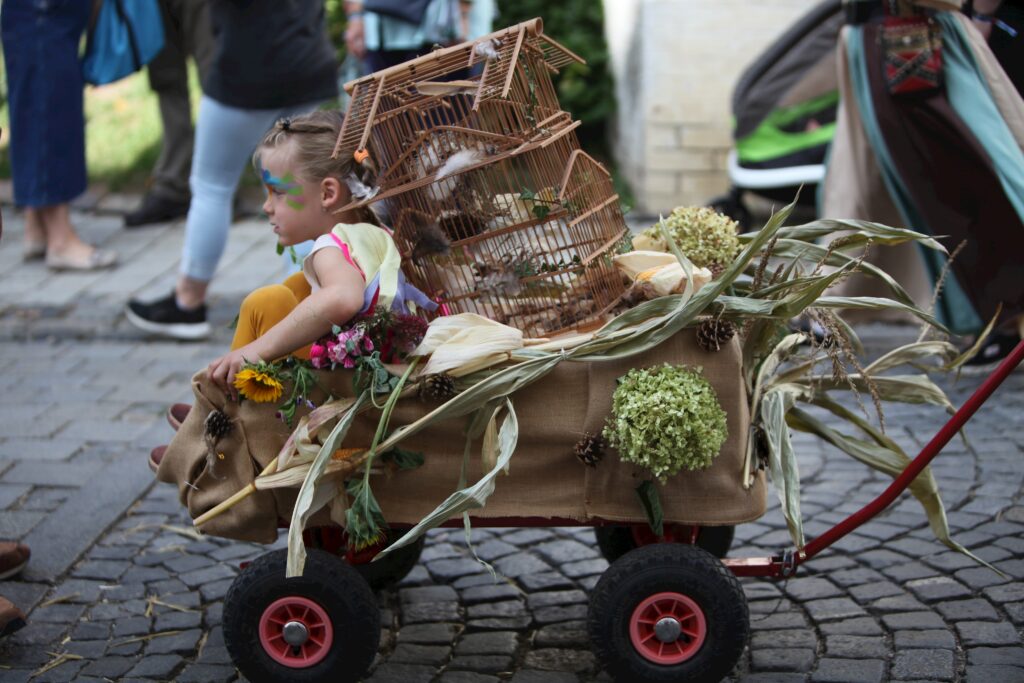Ein Kind in einem Wagen mit Vogelkäfig, Korn und Blumen auf gepflasterten Steinen.