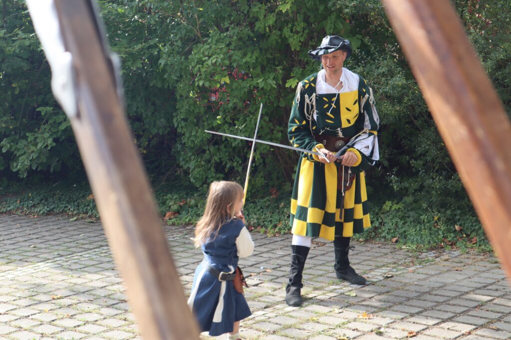 Männlicher Krieger in traditioneller Rüstung und einem Kind mit Schwert auf einer gepflasterten Fläche.