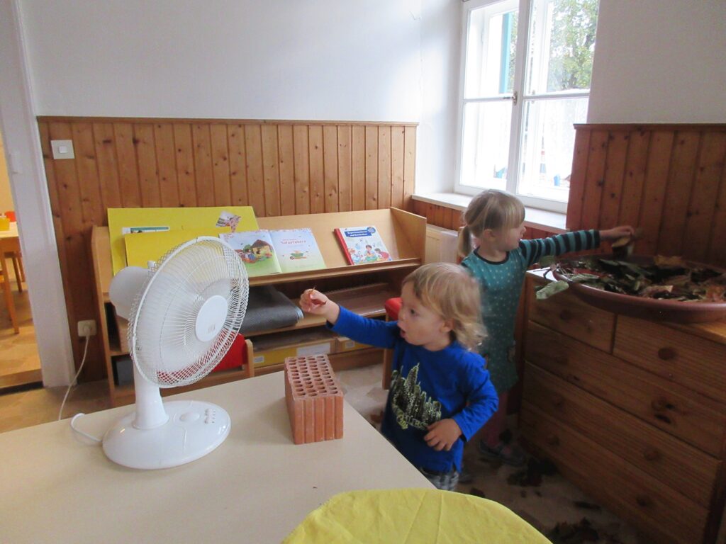 Zwei Kinder in einem Kinderspielzimmer mit Schiebetüren, Kiste, Stuhl, Tisch und Fenster. Ein Kinderzelt.
