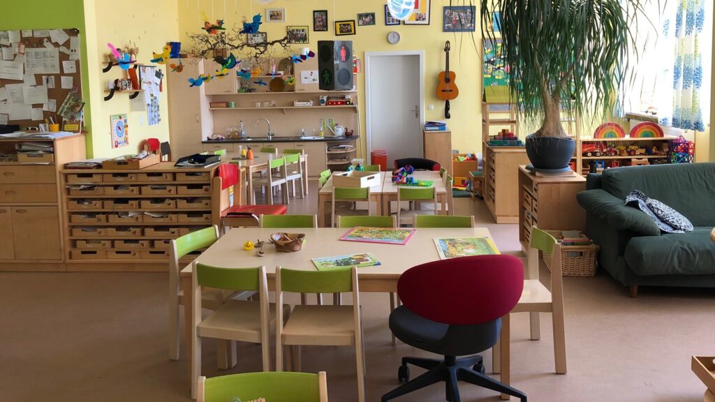 Ein bunt eingerichteter Kindergartenraum mit Spielzeug, Tischen und einem Pflanzenkübel.