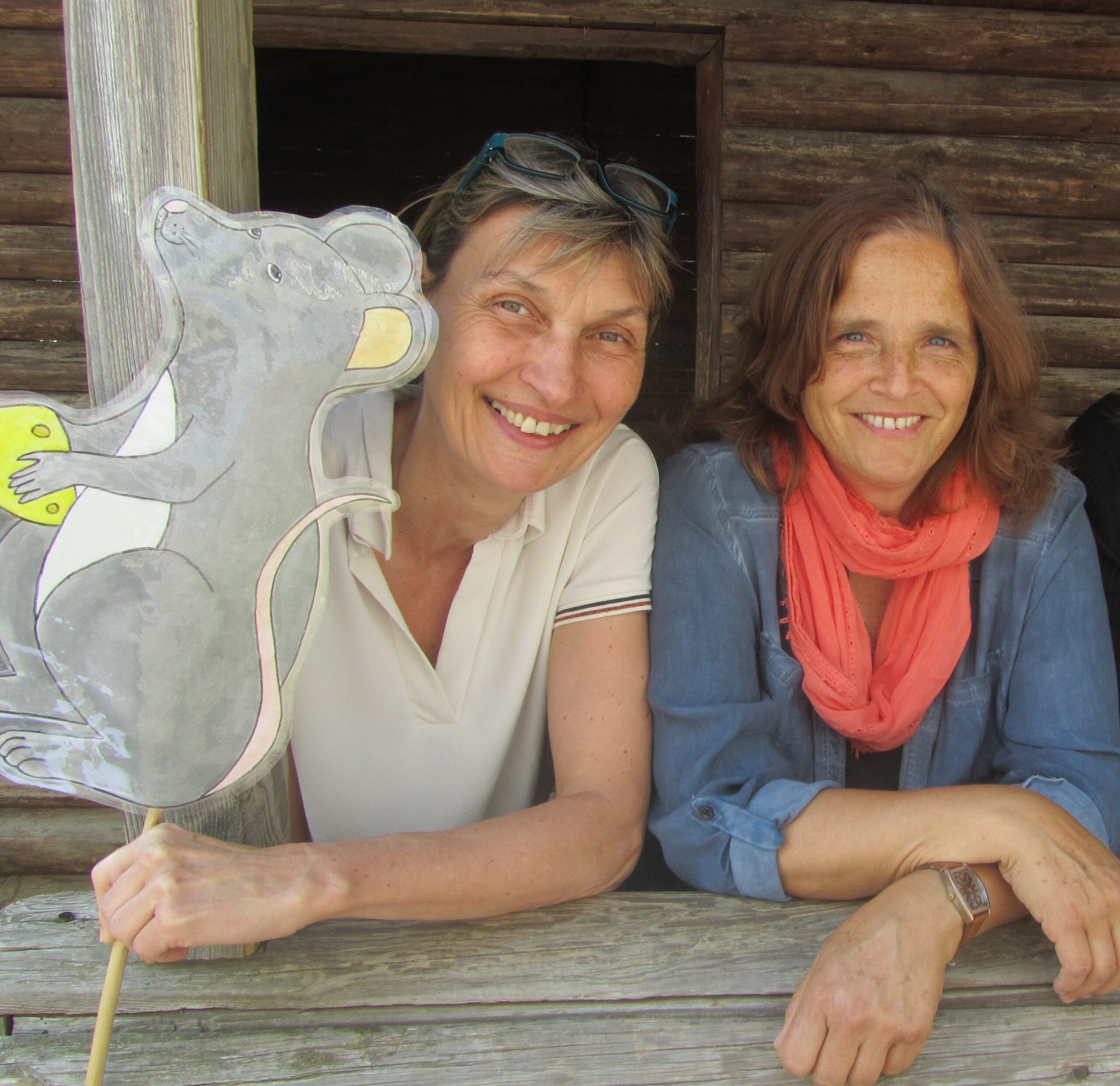 Zwei Frauen mit einer Mäuse-Plakette vor einer Holzstruktur.