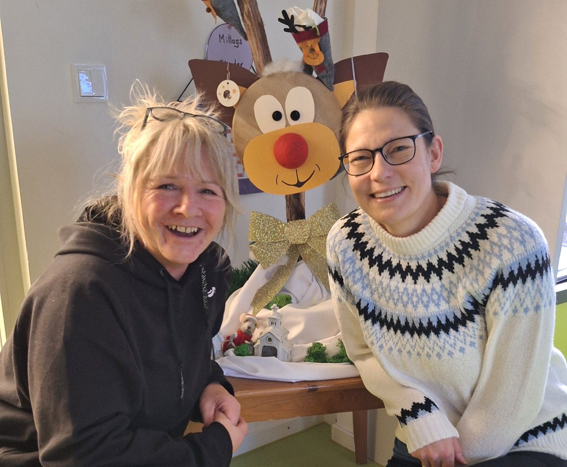 Zwei Frauen lächeln vor einem Weihnachtsdekoration mit Rentier und Hausmodell.