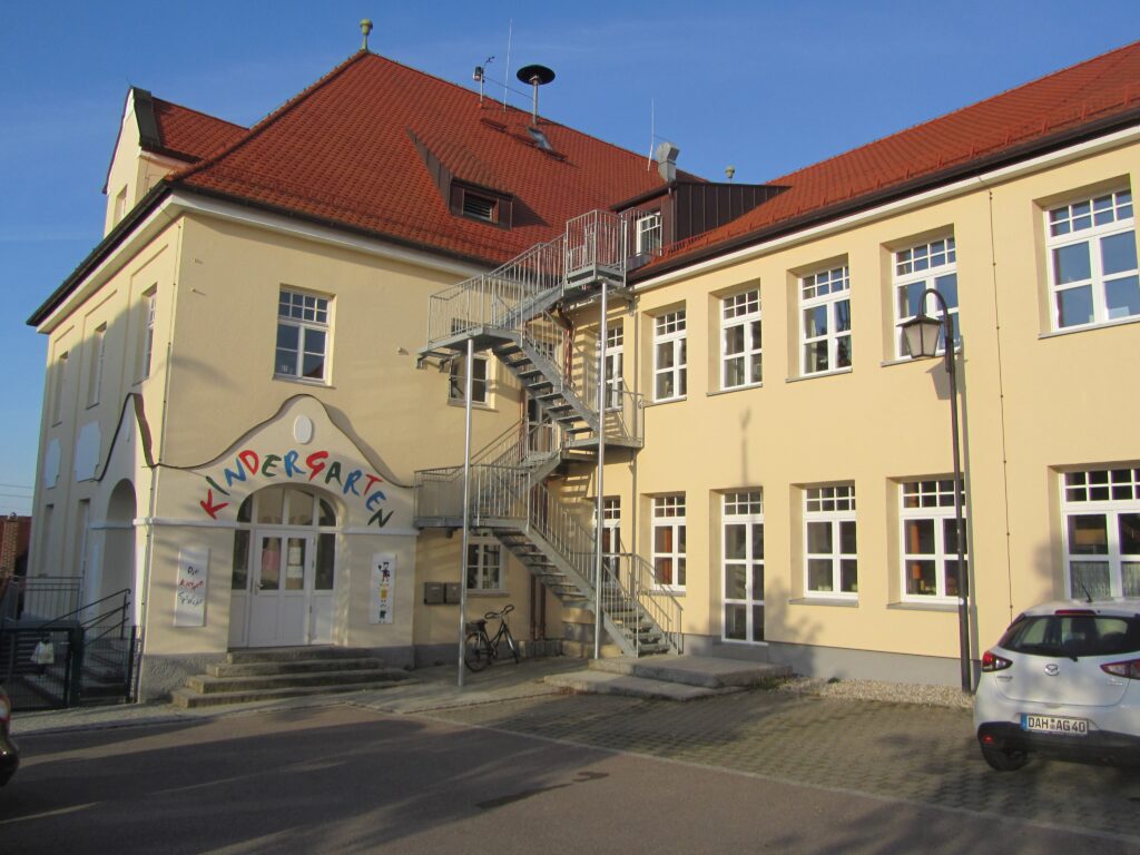 Kindergarten in Altomünster, gelb gestrichen mit roten Schindeln, Schutzschild mit Ausgangstür und Aufgang.