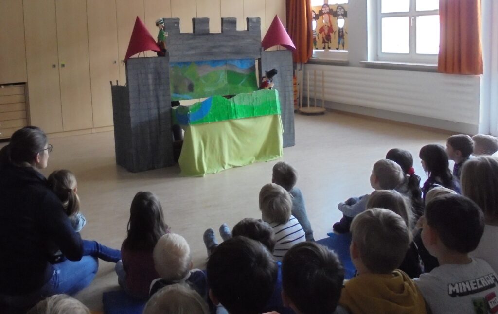 Kinder schauen auf Bühne mit Papierbuden und Puppen in einem Schulraum.