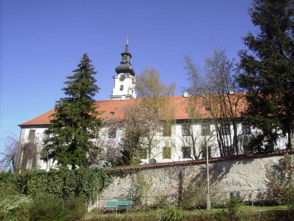 Baudenkmal mit Turm und weißer Fassade, umgeben von Bäumen und Zaun.