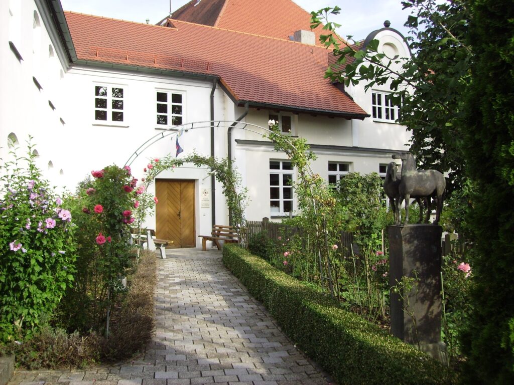 Markt Altomünster, gepflegte Gärten mit Pflanzen, Stele und Weg zu der Eingangstür.
