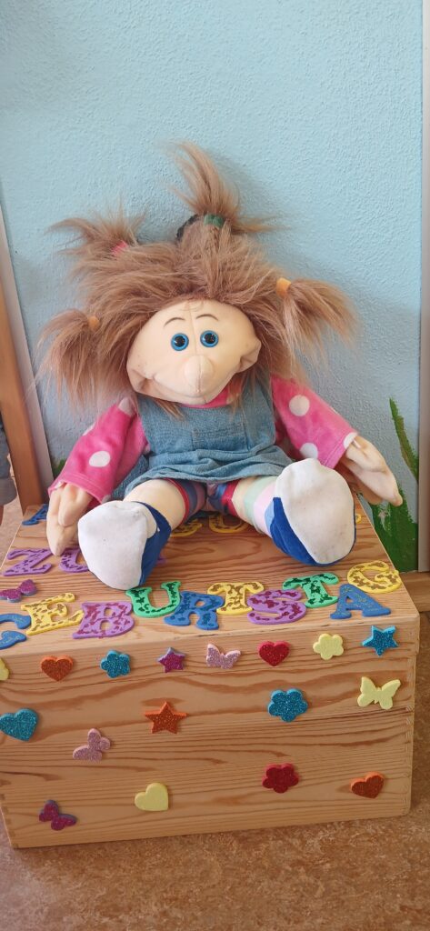 Plüschpuppe mit rosa Kleidung, auf einem Holztisch mit bunten Buchstaben und Figuren.
