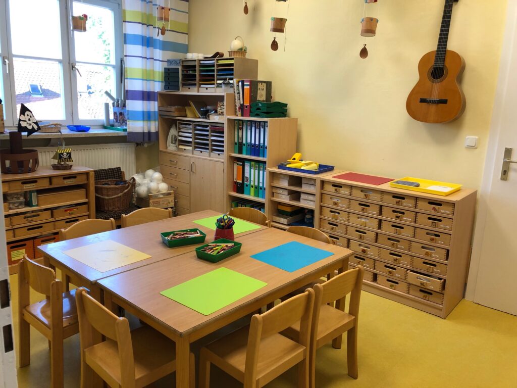 Klassenzimmer mit Tisch, Stühlen, Schreibtisch und Koffer.
