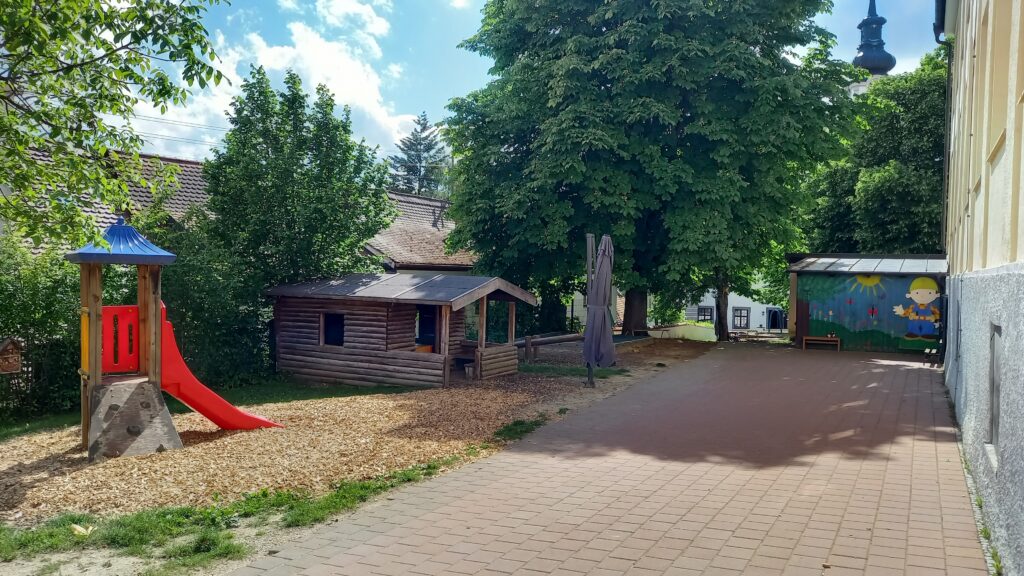 Kinderspielplatz mit Kletterbaum, Schaukel und Holzhaus im Sonnenschein.