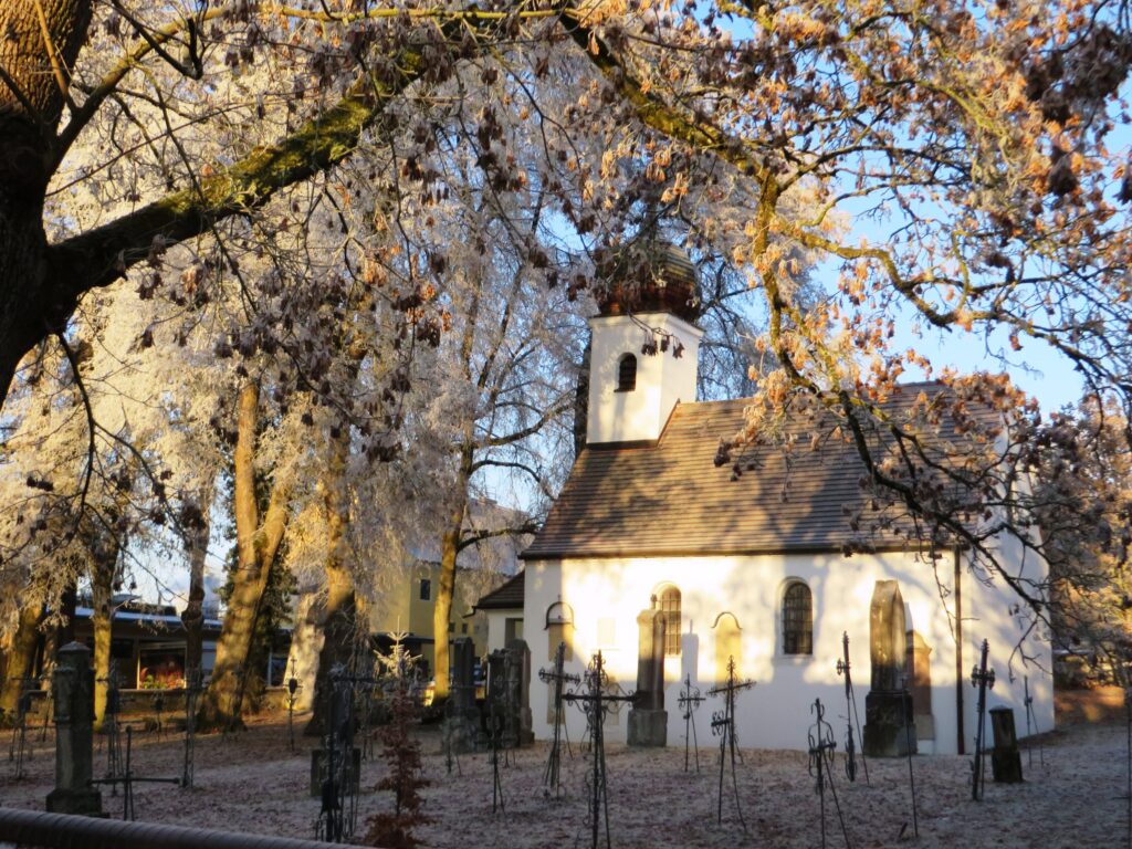 weißes Kirchengebäude im Friedhof mit Frost auf den Bäumen und Tannen im Winter.