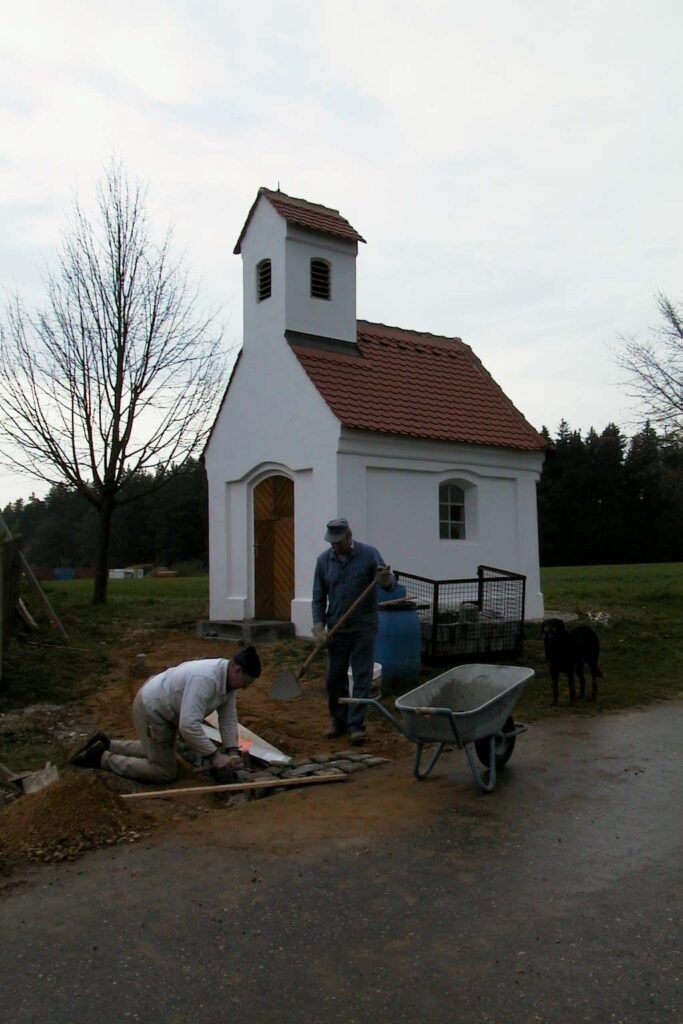 Ein kleines weißes Gotteshaus mit rotem Dach, in dem zwei Männer bei der Ausarbeitung der Umgebung arbeiten.