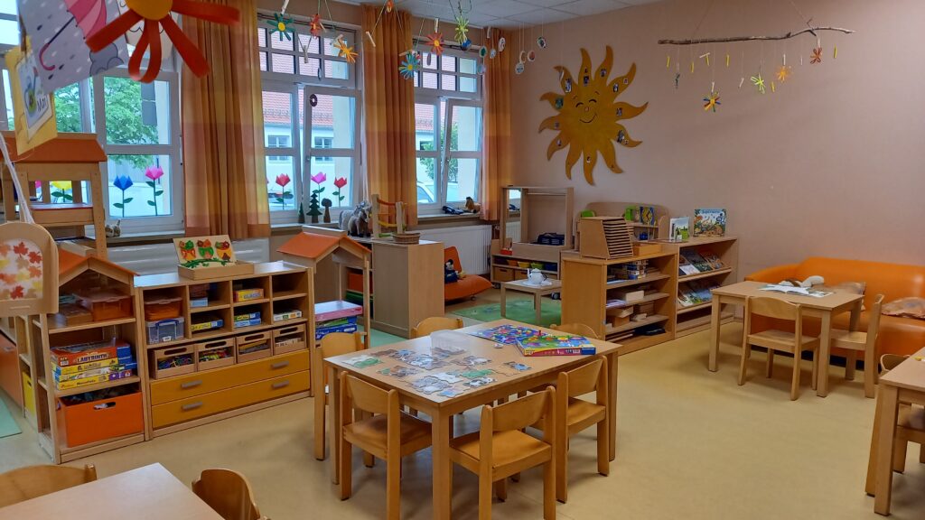 Kindergartenraum mit Tische, Stühlen, Spielzeug und einer großen Sonne an der Wand.